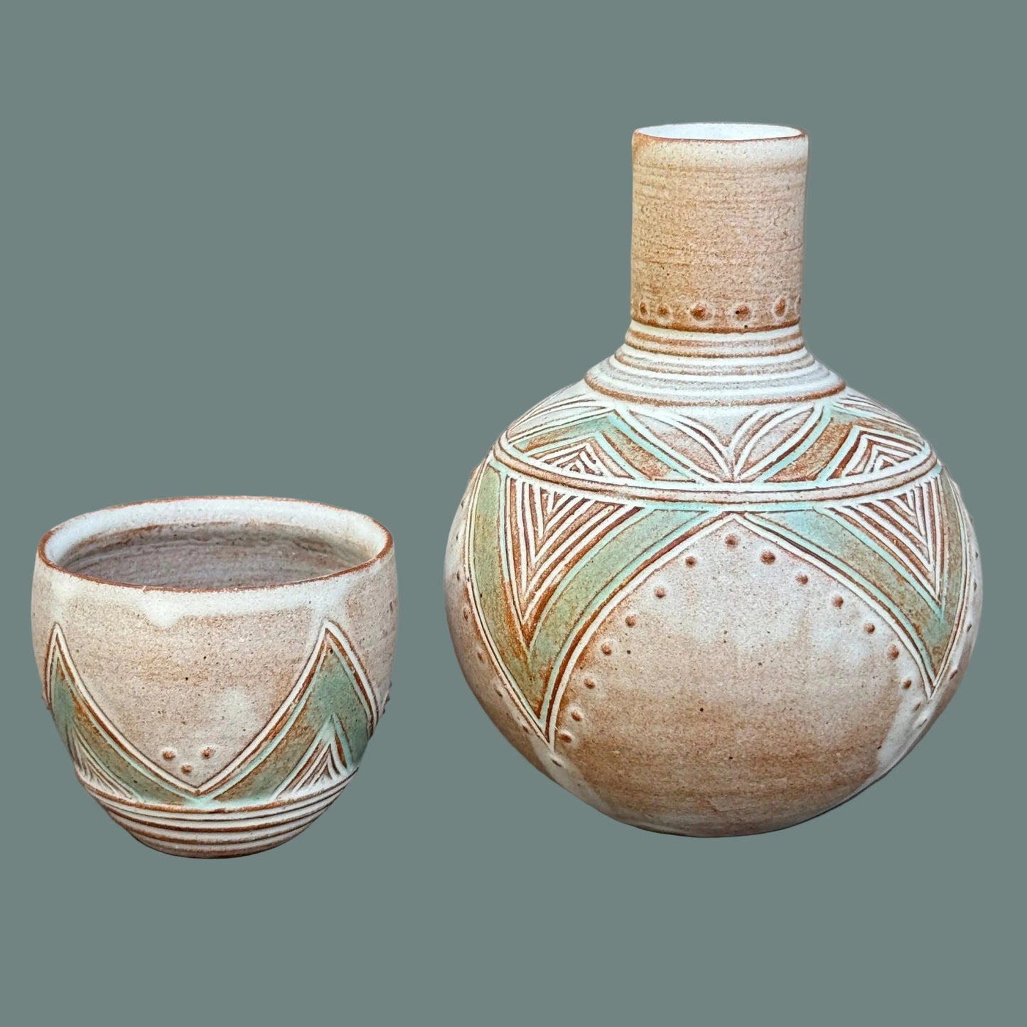 A ceramic set featuring a small cup and a larger rounded vase with a narrow neck, both decorated with earthy brown geometric patterns and light green accents on a speckled beige glaze, set against a dark gray background.