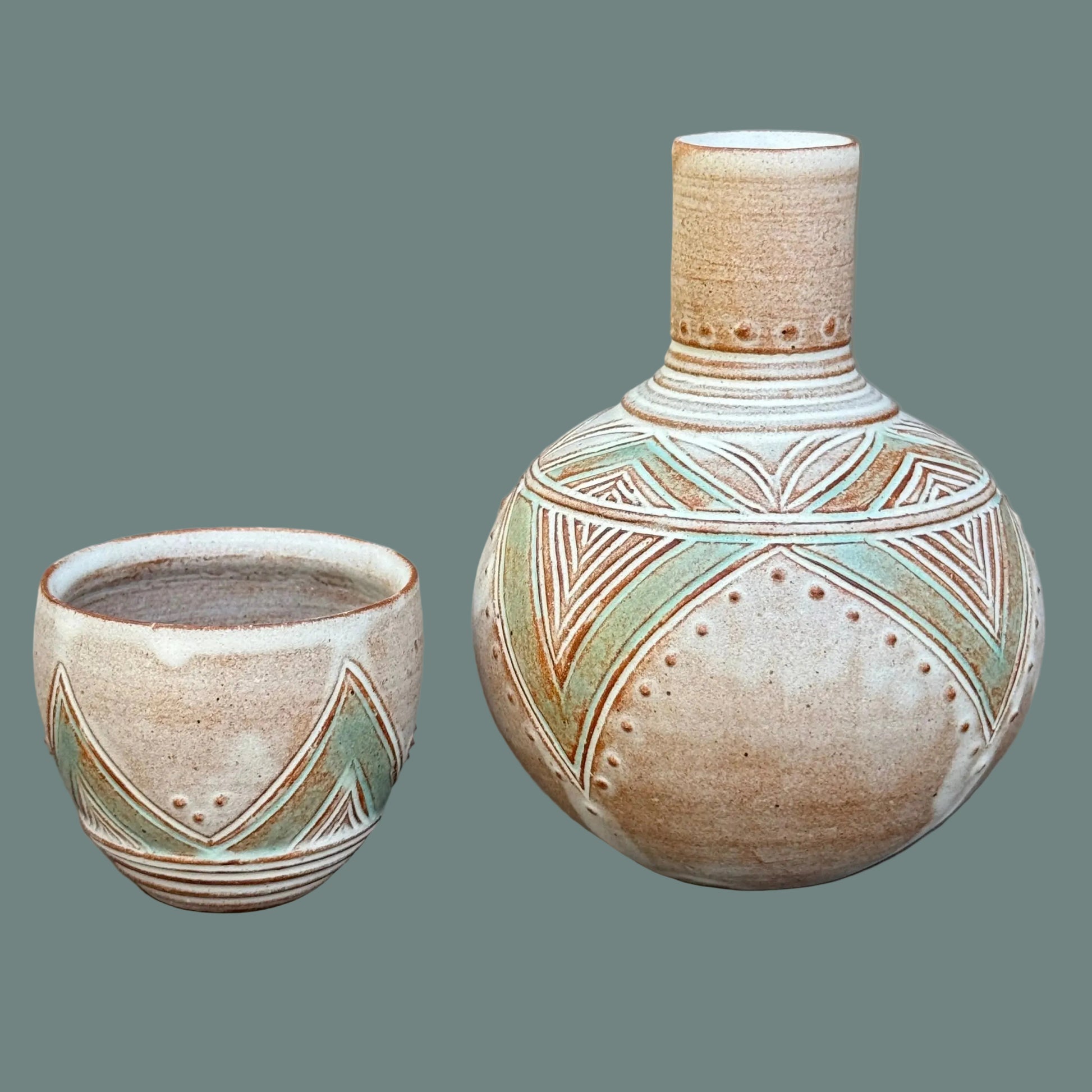 A ceramic set featuring a small cup and a larger rounded vase with a narrow neck, both decorated with earthy brown geometric patterns and light green accents on a speckled beige glaze, set against a dark gray background.