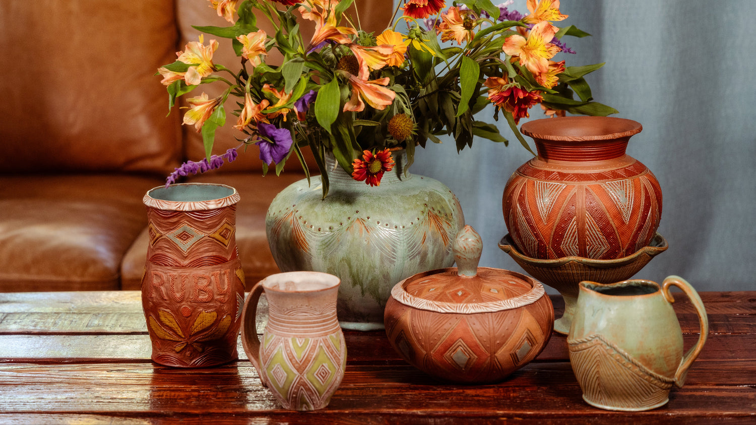 A close shot of a group of pottery on a wood coffee table with a tan leather sofa and light blue curtain in the back. The largest piece of pottery in the photo is an aqua color vase with an unruly wildflower bouquet. The colors of the pottery are earthy and rich, and the surfaces feature rhythmic geometric patterns.